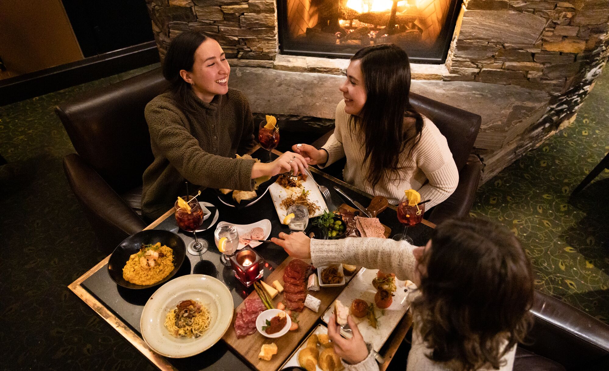 Three ladies enjoy a meal in Banff National Park. There are cheeses, meats, bread and wine on the table as seen from above and a fireplace behind them.